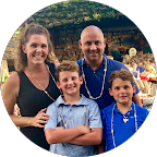 A smiling woman, Aimee Mascioli, stands with a man and two young boys in a casual family photo. They are all wearing beaded necklaces.