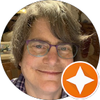 A close-up portrait of Jennifer Haines smiling. She has gray hair and is wearing purple glasses and a plaid shirt. An orange star overlay is in the corner.