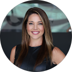 A professional headshot of Sunshine Glover, smiling warmly. She has long brown hair and wears a black top, standing in front of a dark, blurred background.