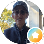 A woman wearing a baseball cap and sunglasses smiles brightly in the sunshine against a background of green trees.